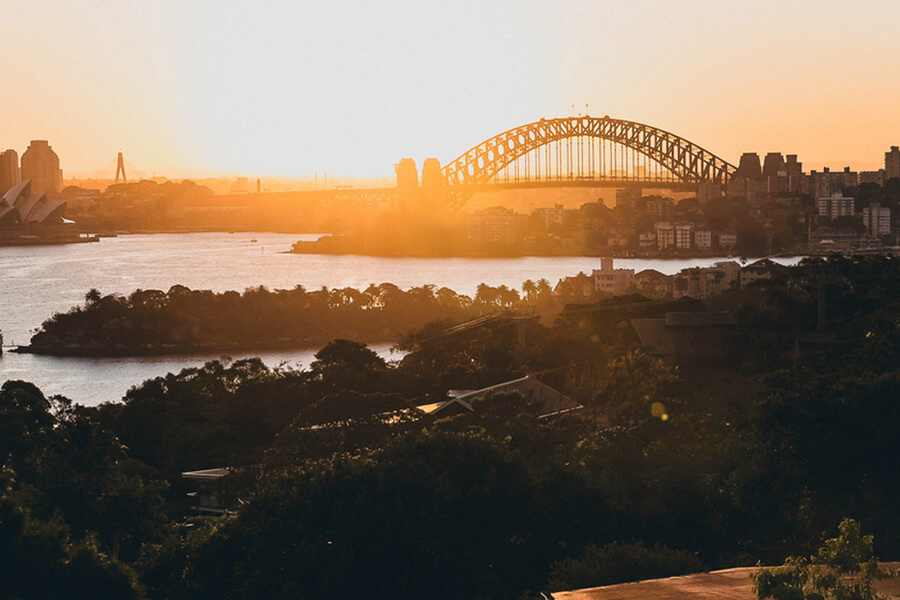 The sun sets over a river with a bridge in the background, surrounded by greenery and city buildings.