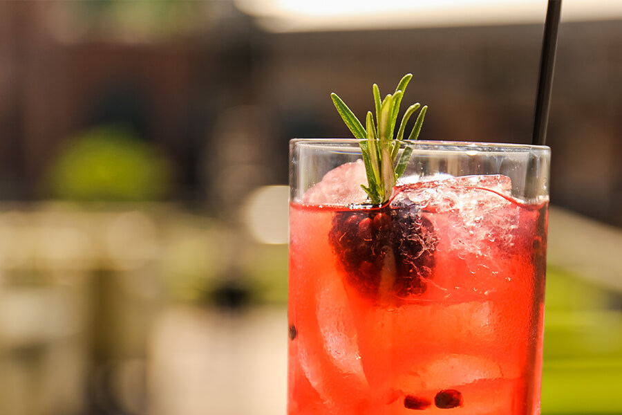 A vibrant red cocktail with ice cubes, garnished with a sprig of rosemary and a dark berry, sits in a clear glass.