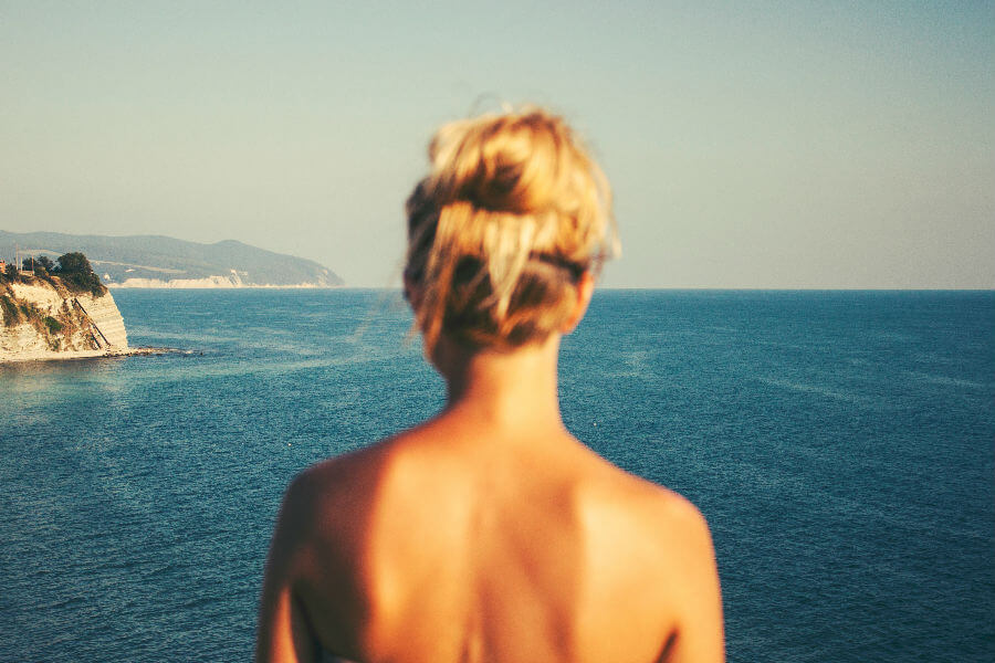 A person with blonde hair in a bun stands by the sea, gazing towards the horizon over calm waters. Coastal cliffs are visible in the background.