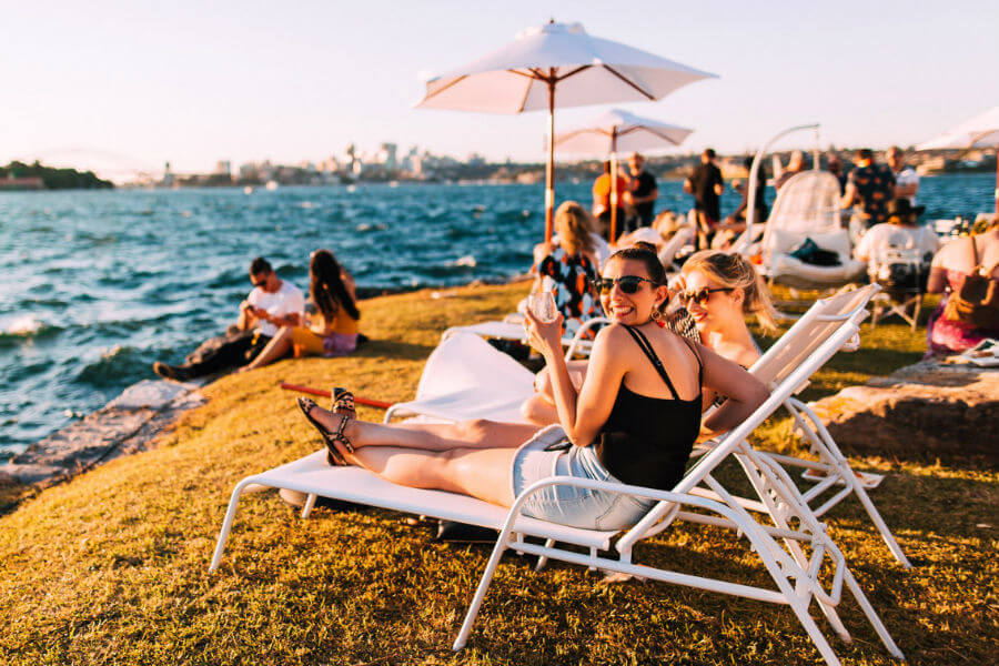 Two women in swimsuits relax on lounge chairs by a waterfront, enjoying drinks, with people socializing and water in the background.