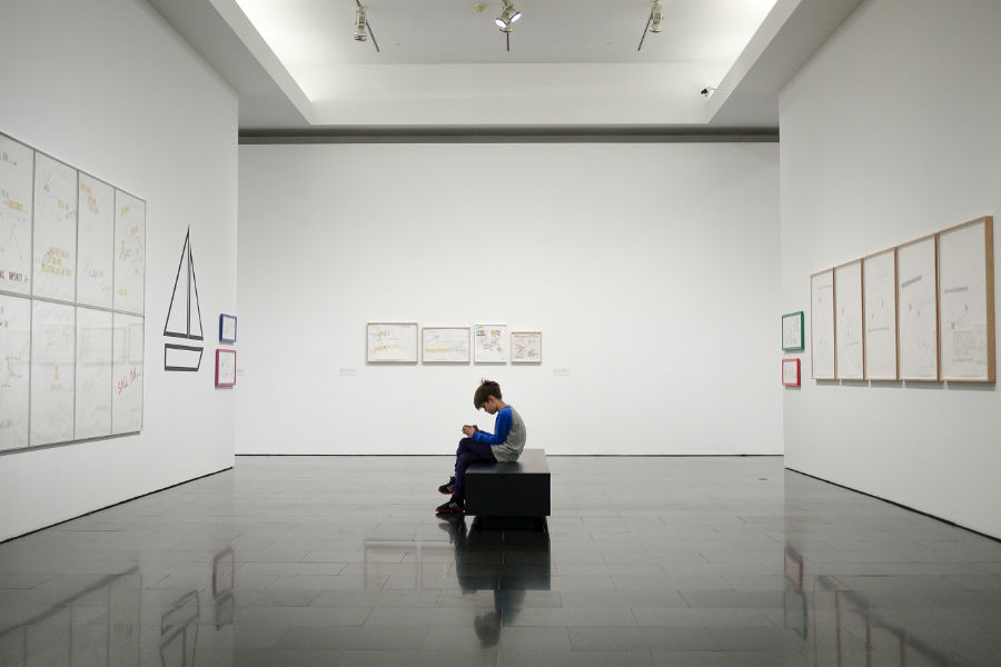 A child sits on a black bench in a modern art gallery, surrounded by white walls and framed drawings.