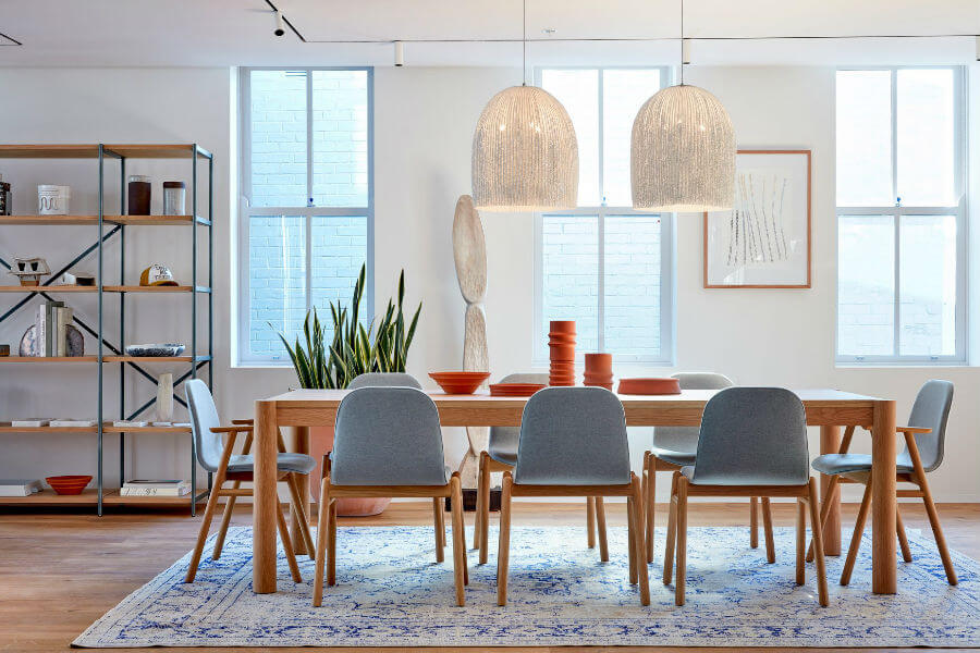 A modern dining area featuring a wooden table surrounded by gray upholstered chairs, with two large pendant lights overhead. A shelving unit displays decorative items, and there are plants near the windows.