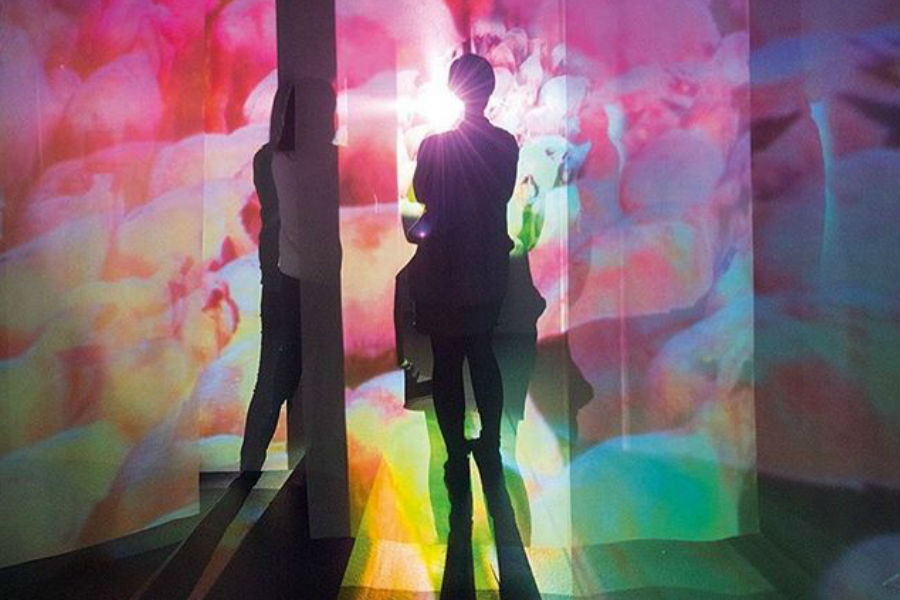 Two silhouetted figures stand in a brightly lit space with colorful projections on the walls.