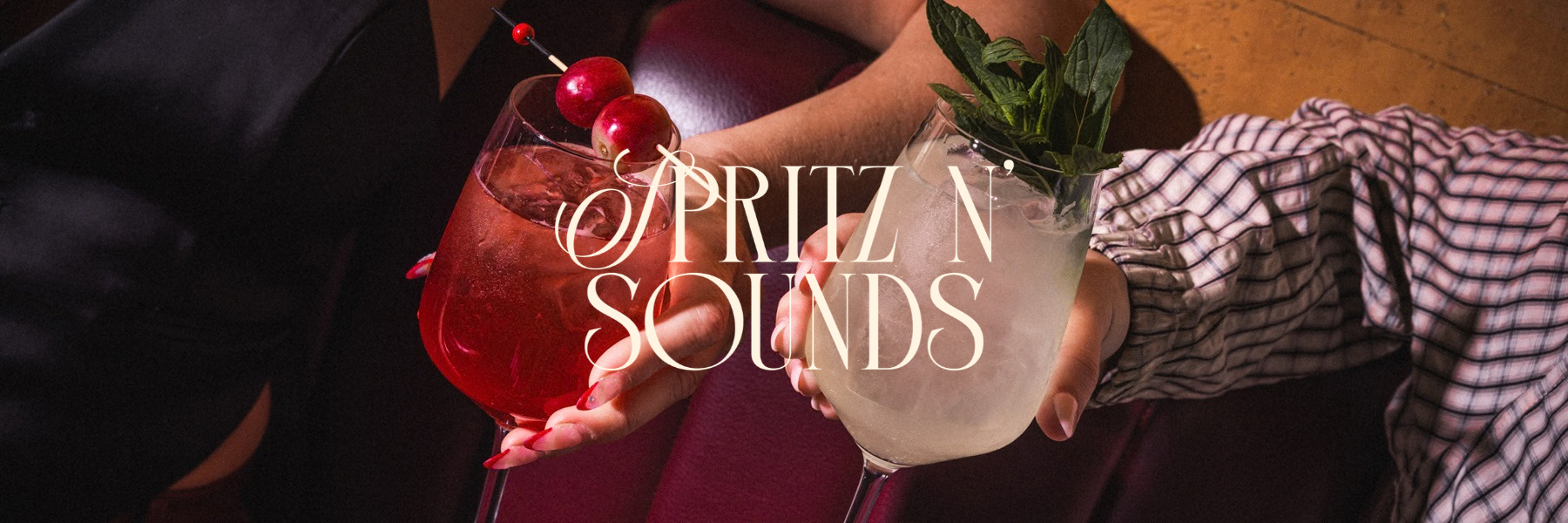 Two hands holding cocktails; one with red drink garnished with cherries and the other with a clear drink topped with mint.