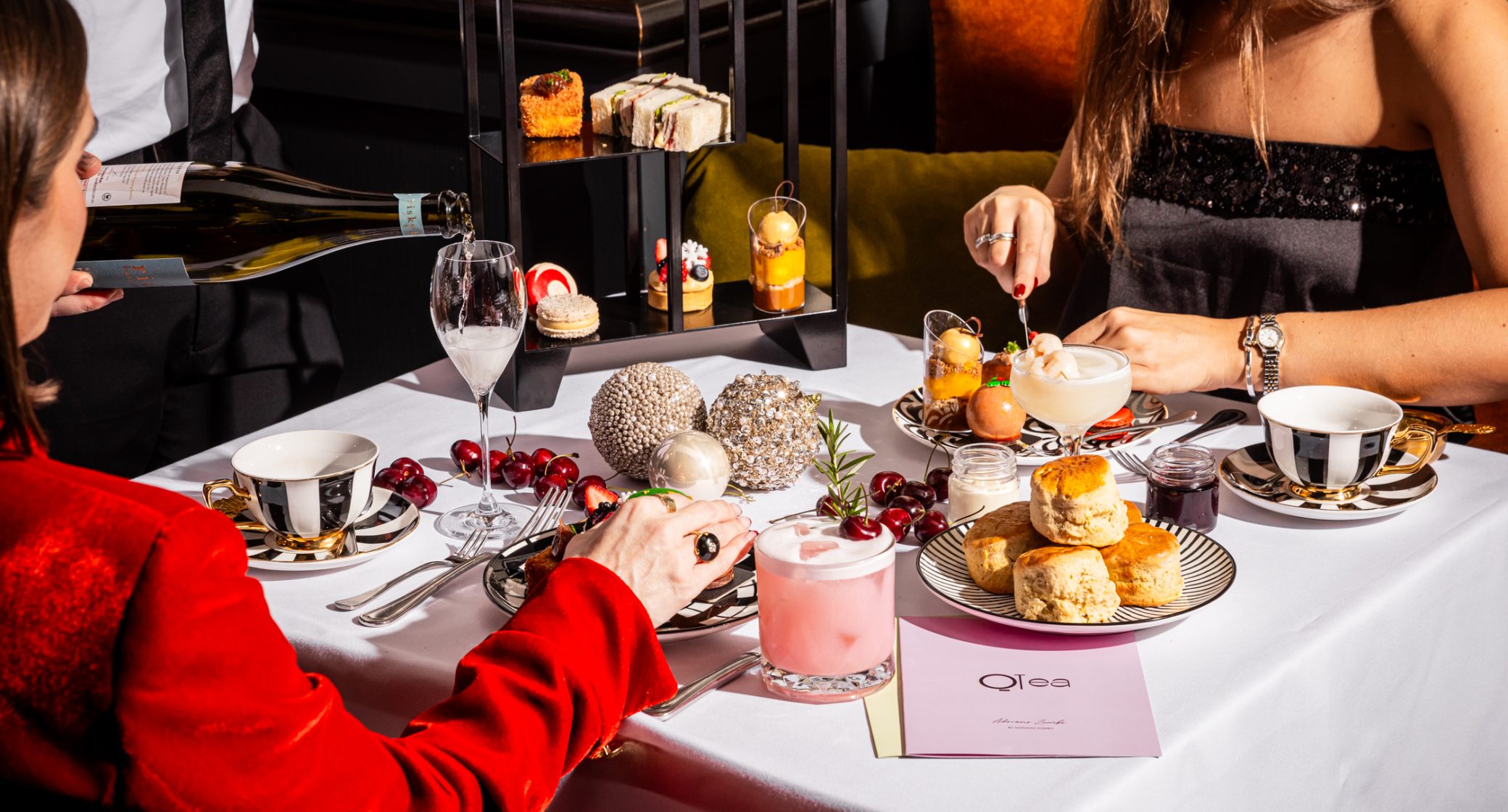 Diners share an elaborate afternoon tea with pastries, scones, fruit, and drinks as wine is poured at the table.