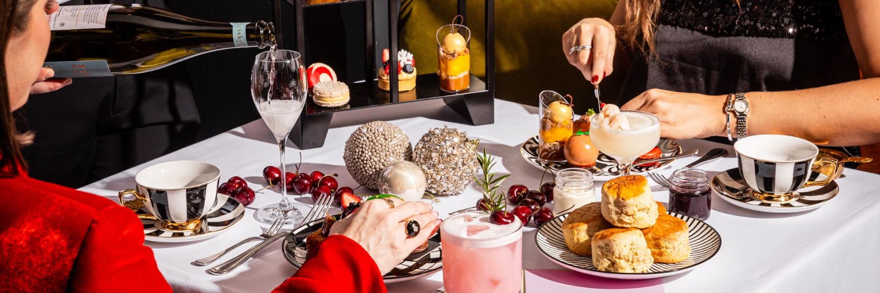 Guests enjoy pastries, cherries, cocktails, and tea as sparkling wine is poured at a white table.