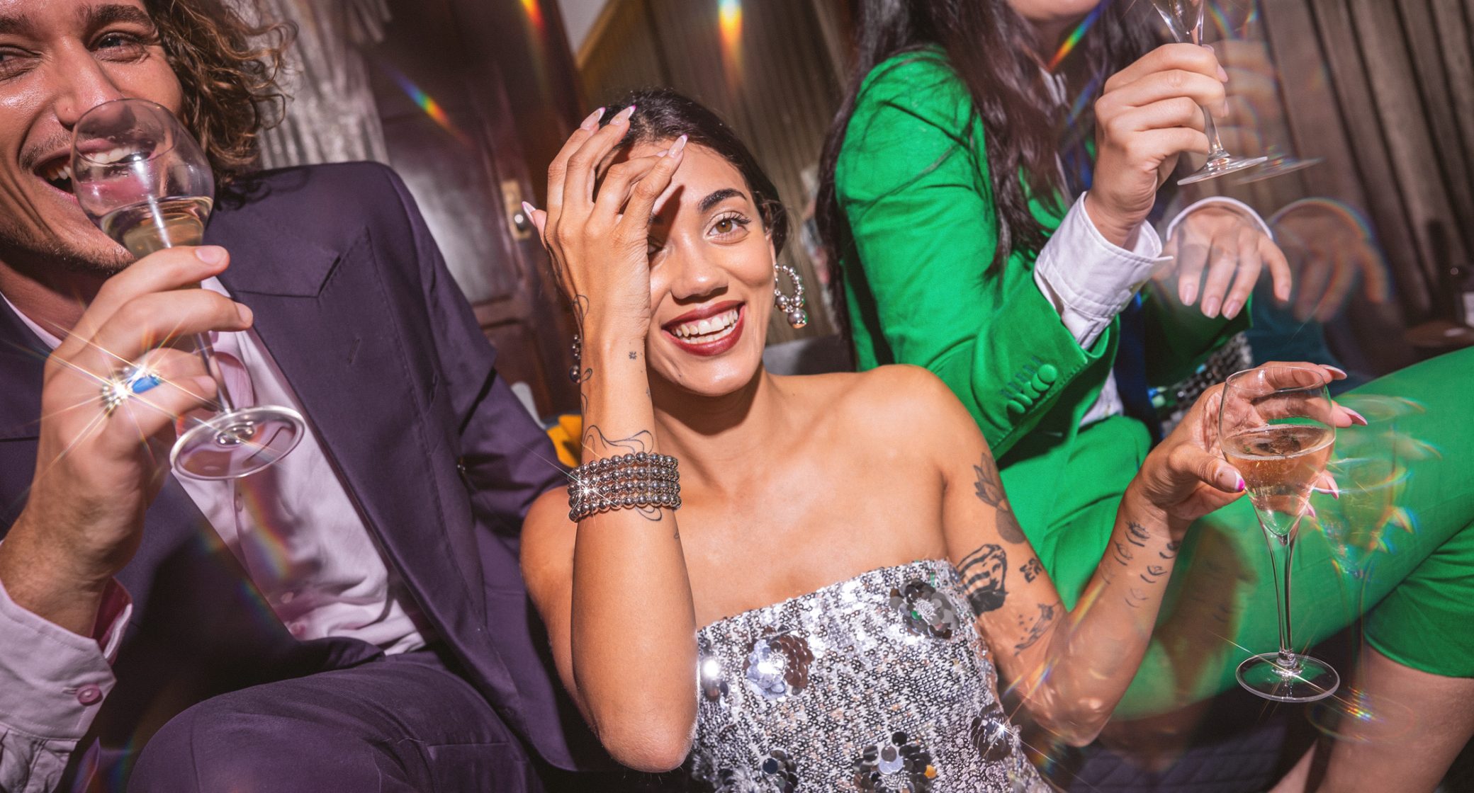 A smiling woman in a sequined dress celebrates with friends, holding wine glasses at a lively gathering.