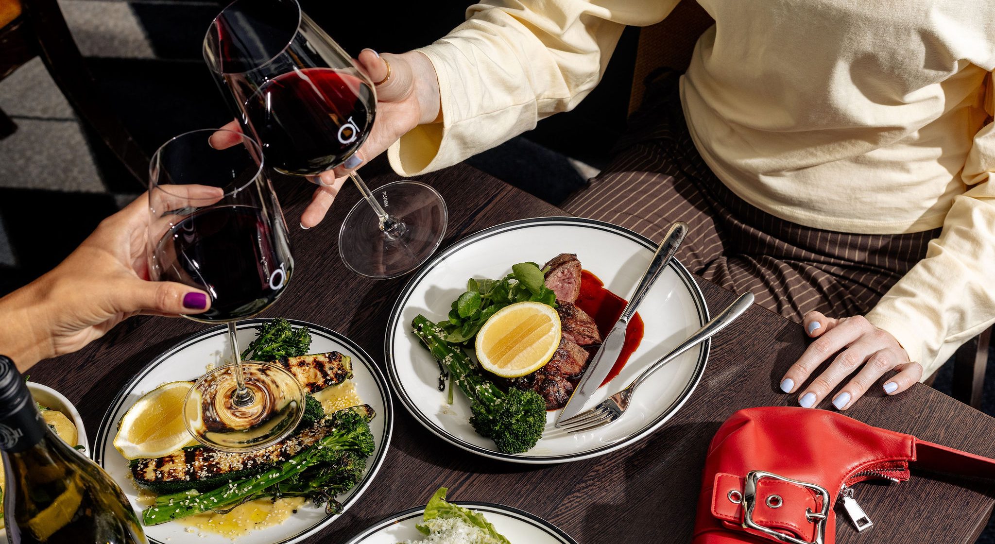 Two diners hold glasses of red wine over a table set with steak, grilled vegetables, and salad.