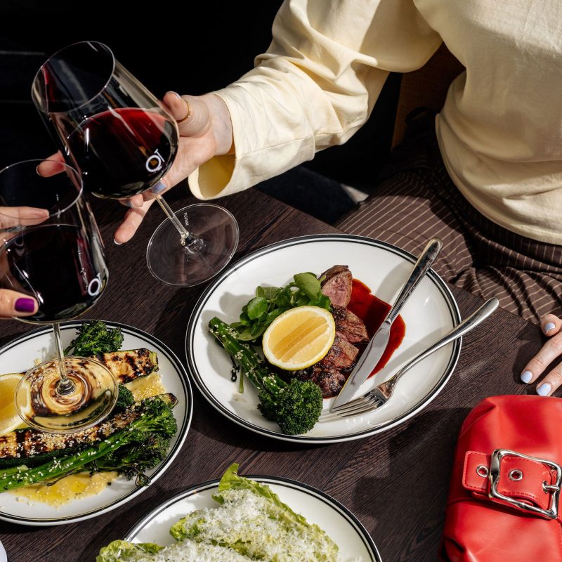 Two diners hold glasses of red wine over a table set with steak, grilled vegetables, and salad.