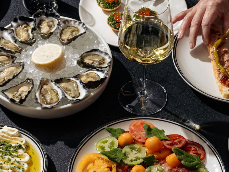 A variety of dishes including oysters, salads, and appetizers are displayed on a table with a wine glass and a red handbag.