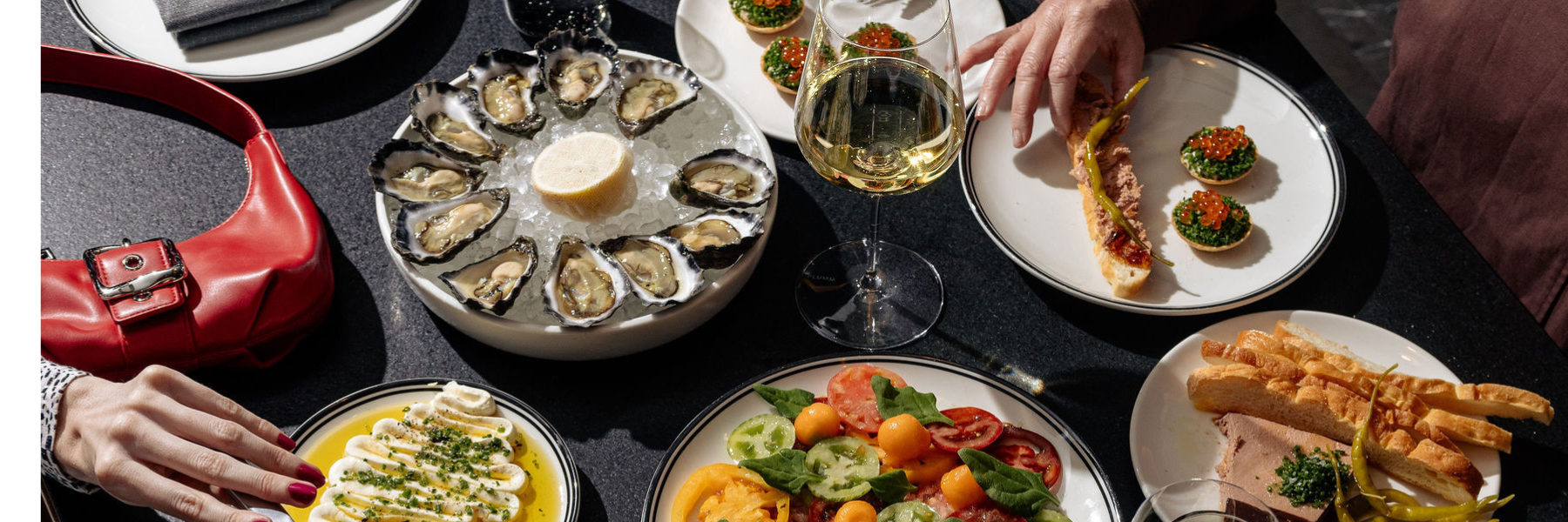 A variety of dishes including oysters, salads, and appetizers are displayed on a table with a wine glass and a red handbag.