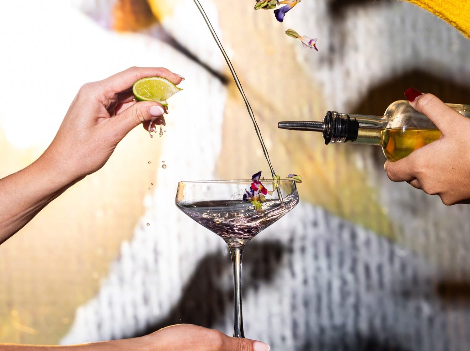 Two hands prepare a cocktail, squeezing lime and pouring liquid while adding flower petals into a glass.