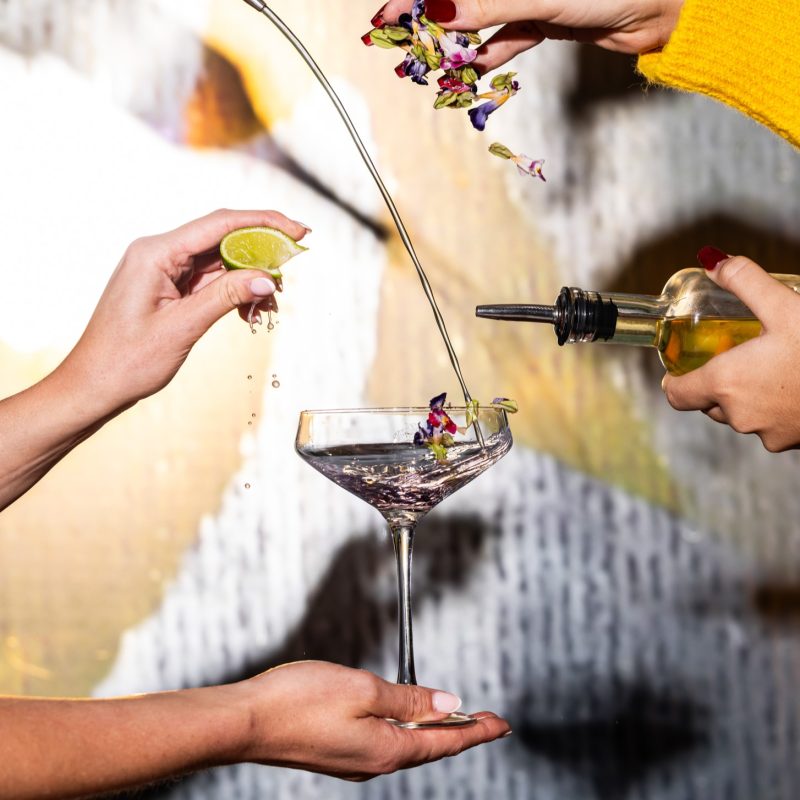 Two hands prepare a cocktail, squeezing lime and pouring liquid while adding flower petals into a glass.