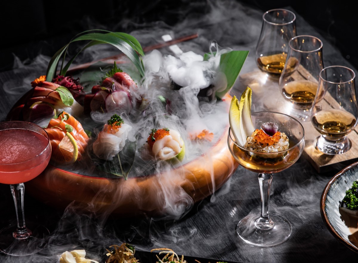 A beautifully arranged platter of sushi and sashimi with smoke, accompanied by cocktails and glasses of whiskey on a dark surface.