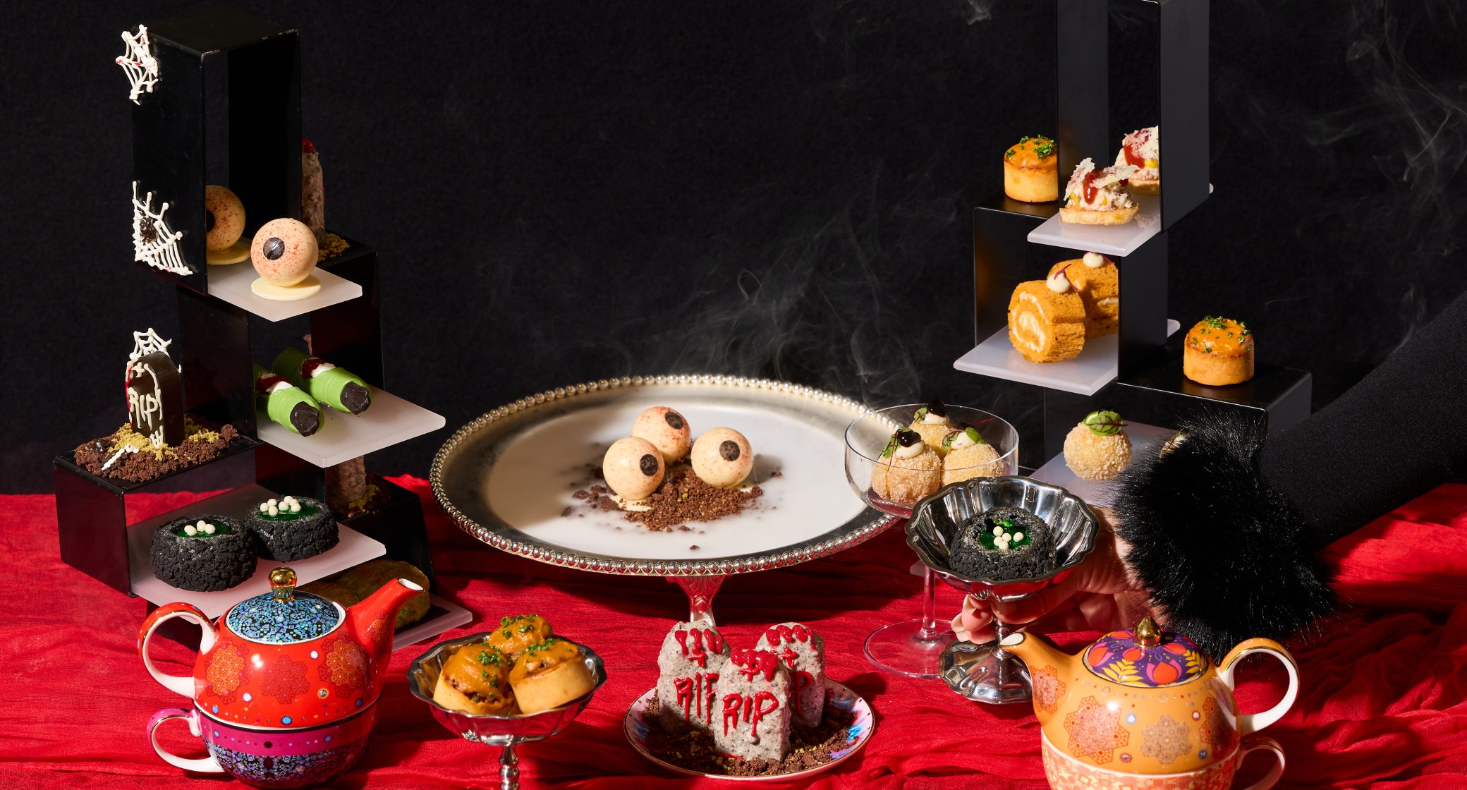 A colorful display of creatively designed desserts on various levels, featuring spooky themes, a silver plate, and decorative teapots on a red fabric backdrop.