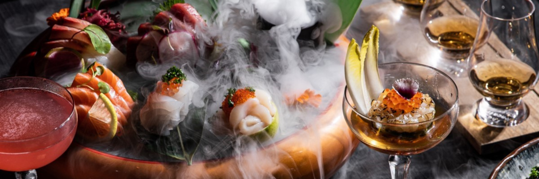 A seafood platter with salmon, scallops, and roe surrounded by swirling vapor, beside a red drink and glasses of amber liquor.