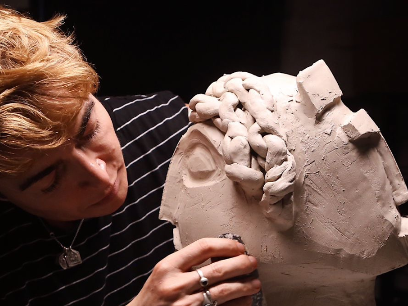 A person leans close to a large clay head sculpture, shaping its surface with their fingers.