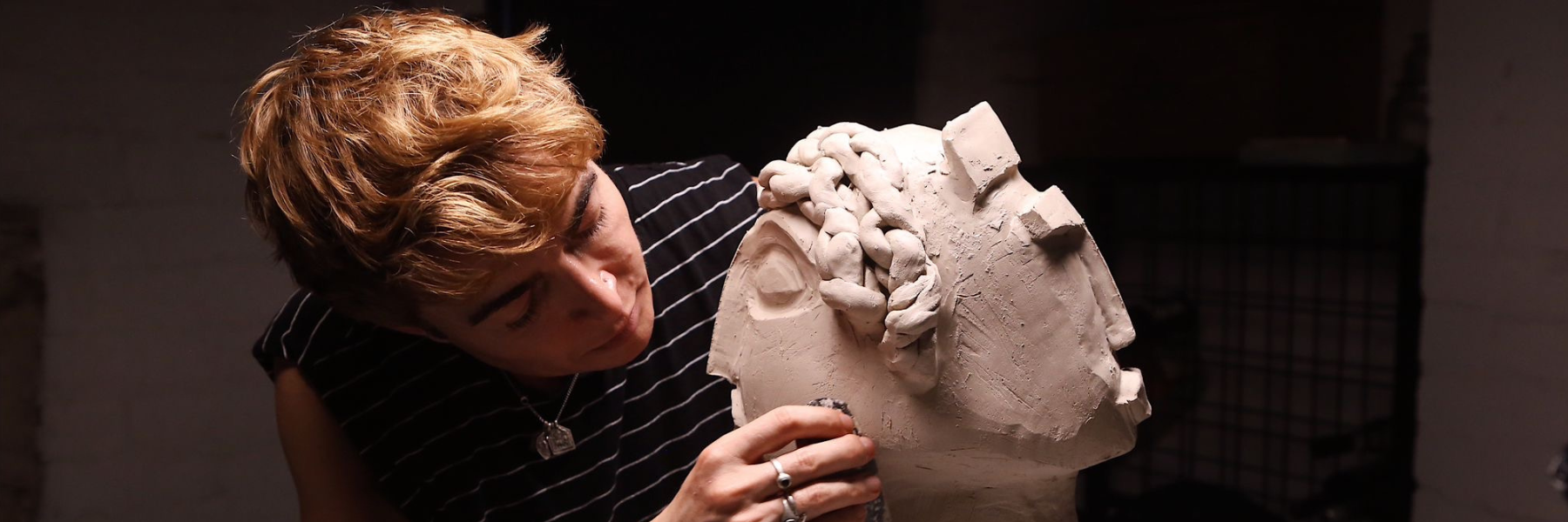 A person leans close to a large clay head sculpture, shaping its surface with their fingers.