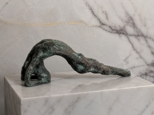 A rough, dark green sculpture arches over a marble pedestal against a veined marble wall.