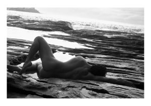 A person lies in shallow water among layered shoreline rocks, one knee bent, in a black-and-white photograph.